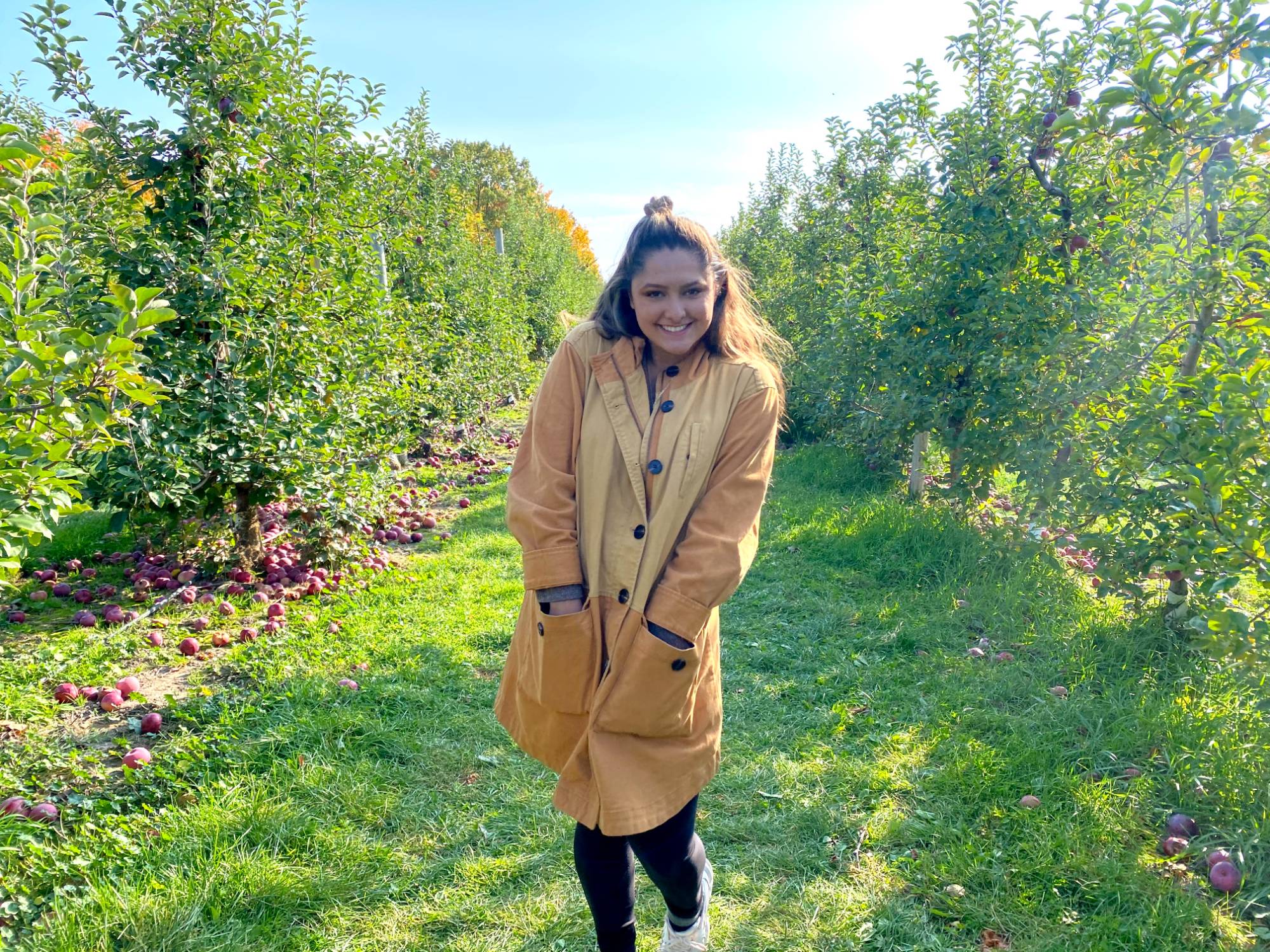 girl with brown hair and a tan coat standing in an apple orchard